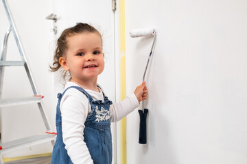 Little girl painting the living room wall with a roller, wearing overalls and a white sweatshirt