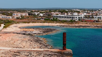 Turquoise waters washing rocky shoreline at es trenc beach, rusty bollard framing resort buildings...