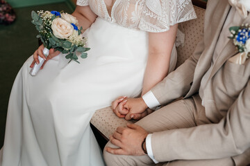 Wedding couple sitting close together, hands gently resting, holding a bouquet.