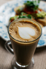 A cup of coffee with a heart on top sits on a table next to a plate of food