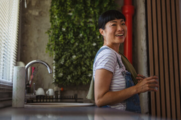 happy mature japanese woman wiping drinking glass at home kitchen