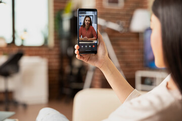 Remote employee sits on sofa, looking at digital device screen during online collaboration with...
