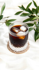 A glass of dark beverage with ice sits on a coaster beside green leaves. Refreshing drink concept