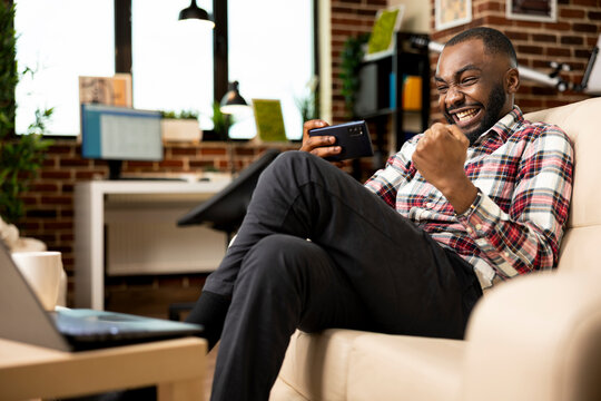 Cheerful male entrepreneur on couch, celebrating with fist raised after winning mobile game. Smiling businessman resting from remote work, holds cellphone, feeling excited from online gaming success.