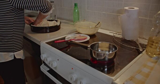 Senior woman preparing dough in her home kitchen. She folds phyllo dough and layers it in a baking pan, getting ready to bake.