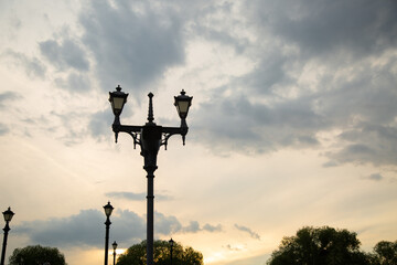 Street black iron design lamp in the park outdoors on summer green trees background under colorful sunset sky