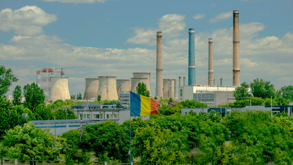 Obraz premium Romanian flag waving in front of a large industrial plant with multiple smokestacks and cooling towers on a partly cloudy day. 