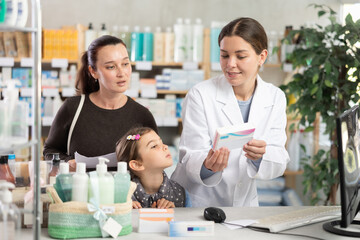Obraz premium Polite young female pharmacist standing by computer showing medicament to mother with daughter in chemistry