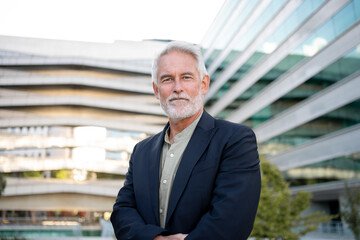 Confident senior businessman in suit standing outside modern glass building. Calm and composed expression in bright urban environment