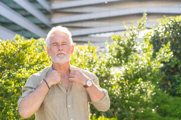 Senior businessman with gray hair standing outdoors in sunlight adjusting shirt collar near modern office surrounded by lush green plants and trees