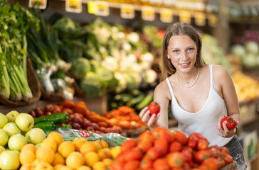 Young woman shopping in vegetable store and buy tomato. Customer carefully examine goods, thinking...