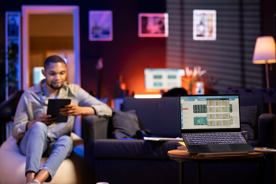 Young Black man relaxes on sofa and uses PC to track forex trades, analyzing stock market graphs for financial growth. Remote worker reviews income trends and trading data for online investments.