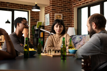 Smiling young woman sits with arms crossed during chess match, calmly observing her male friends...