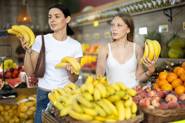 Young and adult women buyers choosing bunch of fresh bananas in vegetable shop