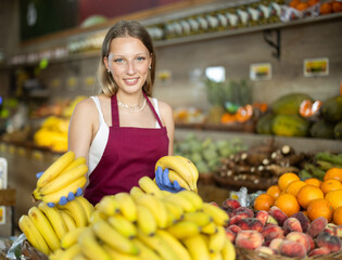 Young woman seller in apron puts bunch of fresh bananas on display in vegetable shop