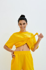 Young smiling woman in yellow casual outfit lifting top, showing toned stomach against plain light...