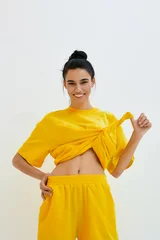Fotobehang Young smiling woman in yellow casual outfit lifting top, showing toned stomach against plain light background, radiating energy and freshness in bright sporty style. © SHOTPRIME STUDIO