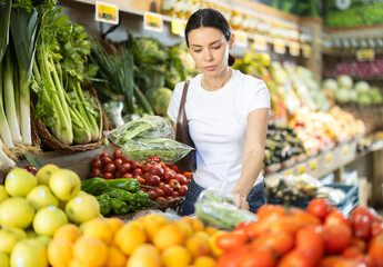 Woman in casual clothes stands near a vegetable counter in a store and chooses edamame. Customer looks at the green beans in the bag and checks them before buying