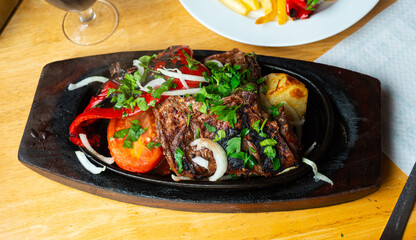Baked pieces of beef with potato and vegetables served in wooden plate decorated with fresh greens and onion