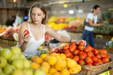 Young blonde woman chooses ripe red tomatoes in the vegetable section of a supermarket. Visitor...