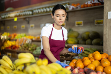 Fototapeta premium Adult woman seller in apron puts fresh peaches on display in vegetable shop
