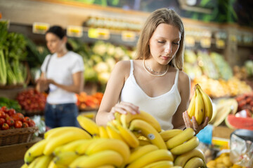 Young woman buyer choosing bunch of fresh bananas in vegetable shop