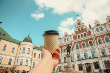 Brown paper cup of natural coffee in women's hand against backdrop of Vyborg Town Hall on sunny day