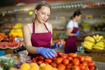 Girl puts ripe tomato in box on display case, arranges assortment in vegetable shop. Worker puts...