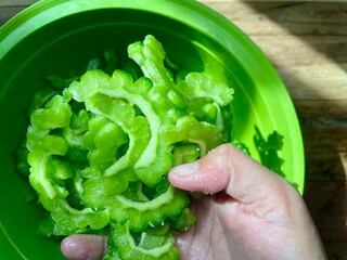 close-up photo of a woman's hand picking up slices of raw bitter melon in a basin on a wooden...