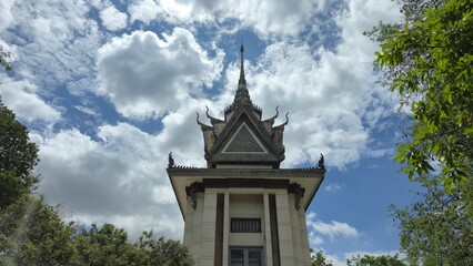 Choeung Ek Killing Fields, Cambodia – 25 June 2025. A white memorial pavilion with tiered carved roof and central spire rises above trees against a cloudy sky.