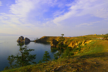 Lake Baikal stretches under a soft blue sky. Calm waters meet green hills and sandy beaches,...