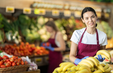Adult woman seller in apron puts bunch of fresh bananas on display in vegetable shop