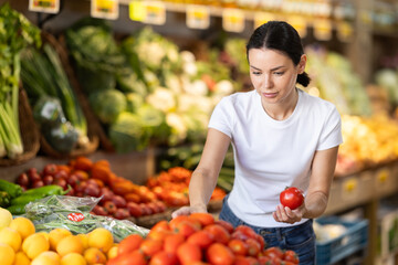 Young woman in casual clothes stands in a supermarket near a vegetable counter and chooses tomatoes. Buying fresh vegetables to cooking