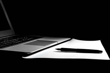 Laptop, pen, and paper on a desk in a monochrome image.