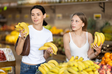 Two women stand near a bountiful fruit stall and pick out ripe bananas at the supermarket. Fruit...