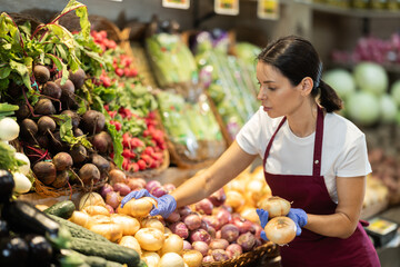 Female vendor in a red apron puts onions on a supermarket window next to other farm vegetables. Seller is busy distributing goods in the vegetable department