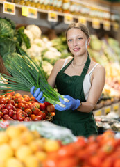 Young woman seller in apron puts green onions on display in vegetable shop