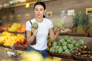 Girl client near vegetable stand in store choose artichoke. Young woman shopping in vegetable...