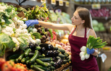 Young woman seller in apron puts green onions on display in vegetable shop