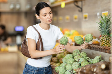Girl client near vegetable stand in store choose artichoke. Young woman shopping in vegetable...