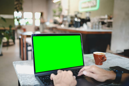 A person typing on a laptop with a blank green screen in a bustling cafe. A versatile chroma key mockup for showcasing websites or video content.