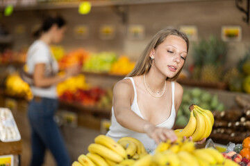 Young woman shopping in vegetable store and buy banana. Customer carefully examine goods, thinking...