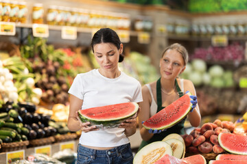 Young woman seller helps adult woman buyer choose fresh watermelon in vegetable shop