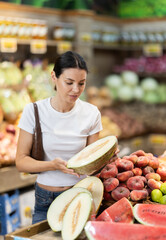 Young woman shopping in vegetable store and buy melon. Customer carefully examine goods, thinking...