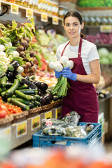 Woman vendor in a red apron is standing near a vegetable counter and laying out shallots. Farm shop worker distributing fresh vegetables