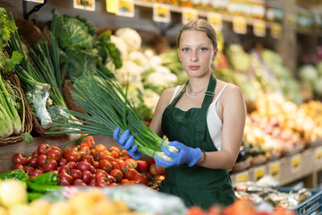 Young woman seller in apron puts green onions on display in vegetable shop