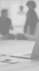 A grayscale image of a laptop and smartphone on a desk, with blurred figures in the background.