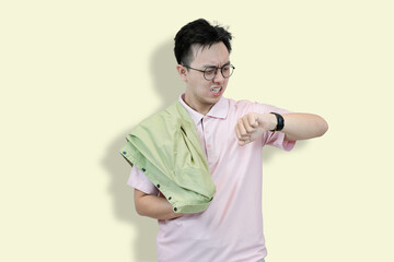 Young Asian man wearing glasses and a polo shirt, holding a jacket, looking at his wristwatch with...