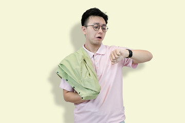 Young Asian man wearing glasses and a polo shirt, holding a jacket, looking at his wristwatch with...