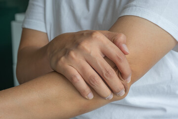 An Asian man sits near a window in soft natural light, supporting their left elbow—suggesting localized joint pain, possible tendon inflammation, or repetitive strain. Healthcare and medical concept.